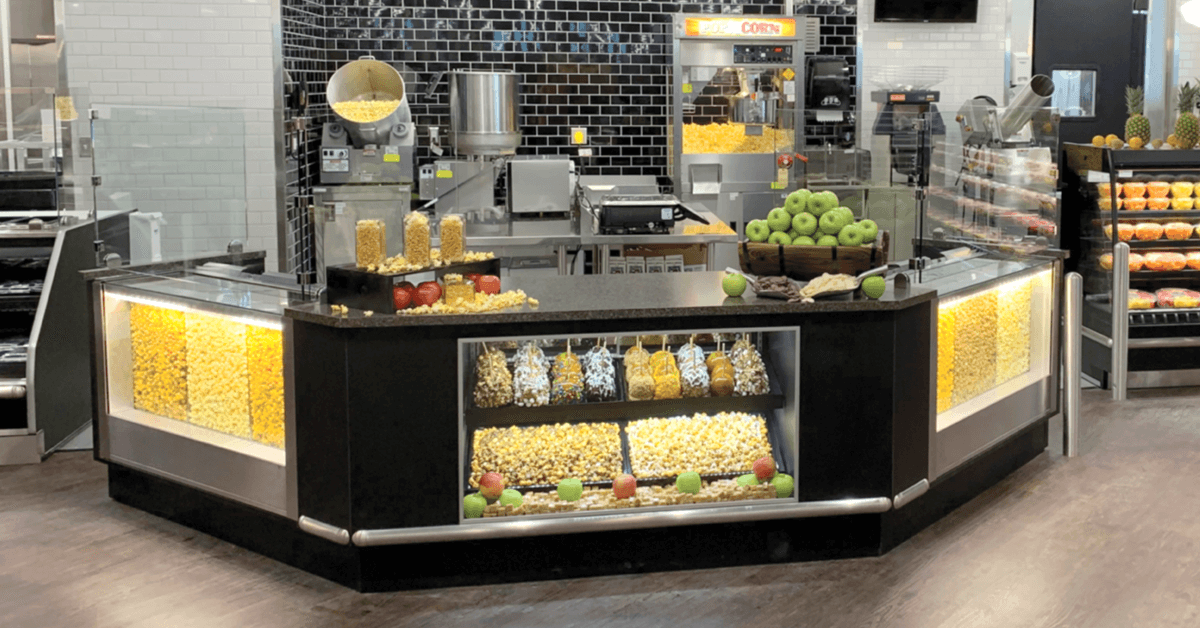 Wide view of a retail food theater concession station featuring popcorn displays, caramel apple dipping equipment, illuminated popcorn bins, and fresh snack merchandising in a grocery-style venue. Wide view of a retail food theater concession station featuring popcorn displays, caramel apple dipping equipment, illuminated popcorn bins, and fresh snack merchandising in a grocery-style venue.